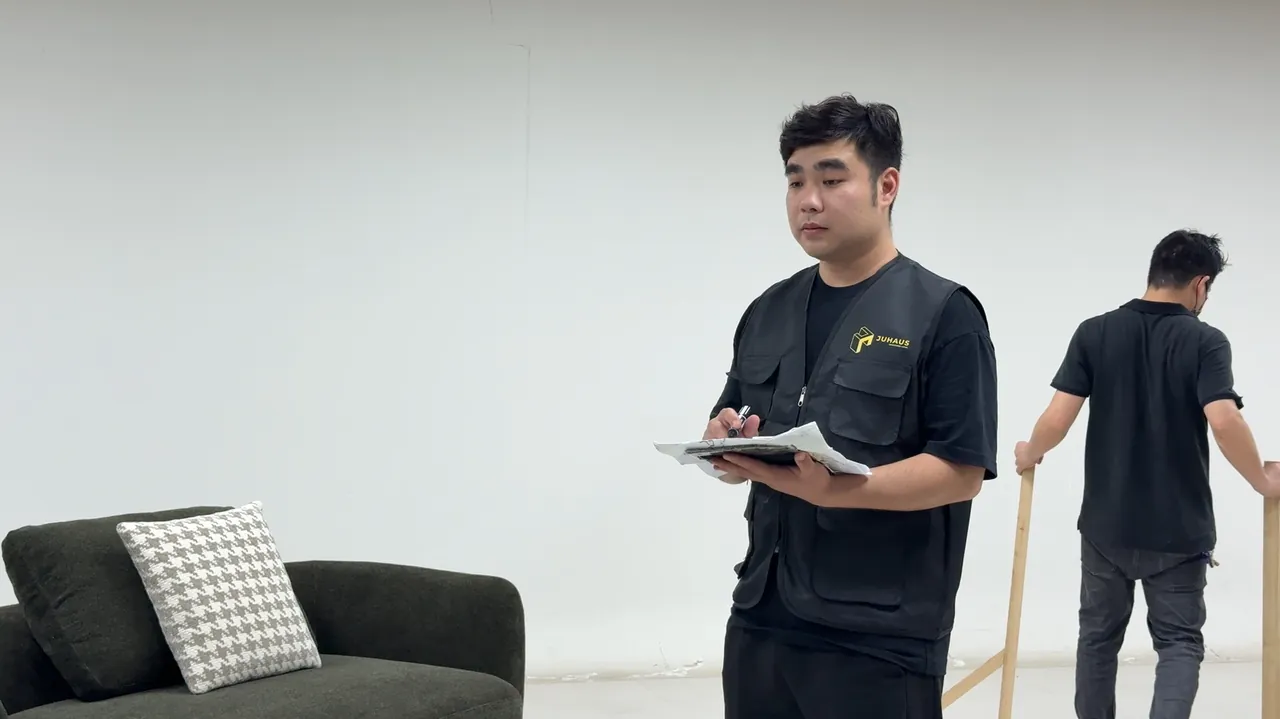 A person in a Juhaus vest holds a clipboard beside a sofa while another person handles a wooden guide.